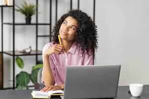Young person sat at a desk with an open laptop, thinking and note taking.